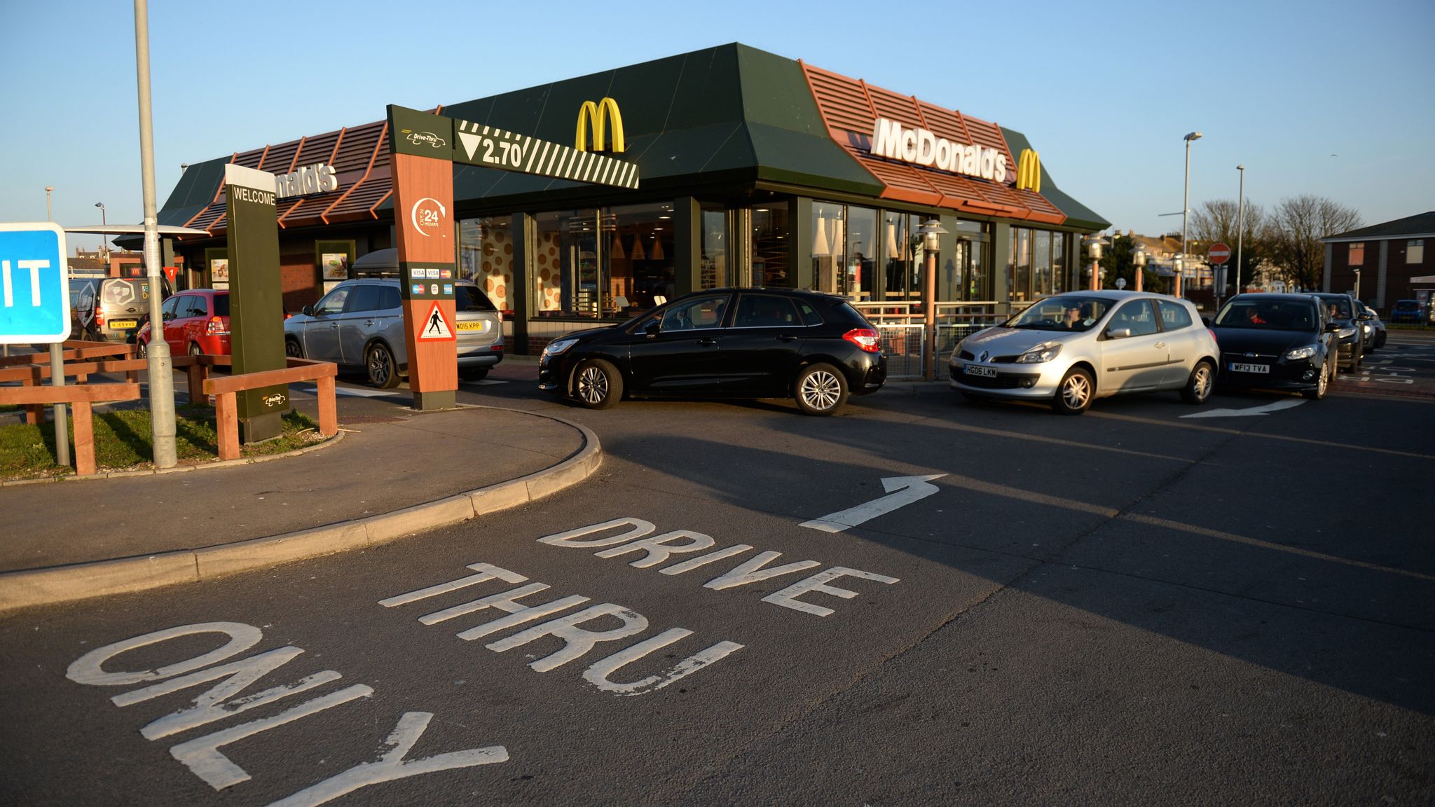 The picture shows the outside of a McDonald's restaurant. Cars are queuing around the side of the restaurant, and on the road in the foreground, it reads "Drive thru only".